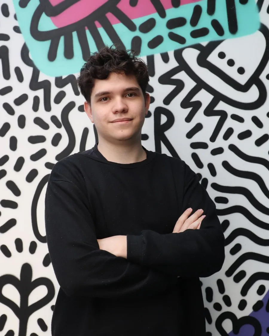 Retrato de Thiago Miedes de braços cruzados diante de um mural grafitado